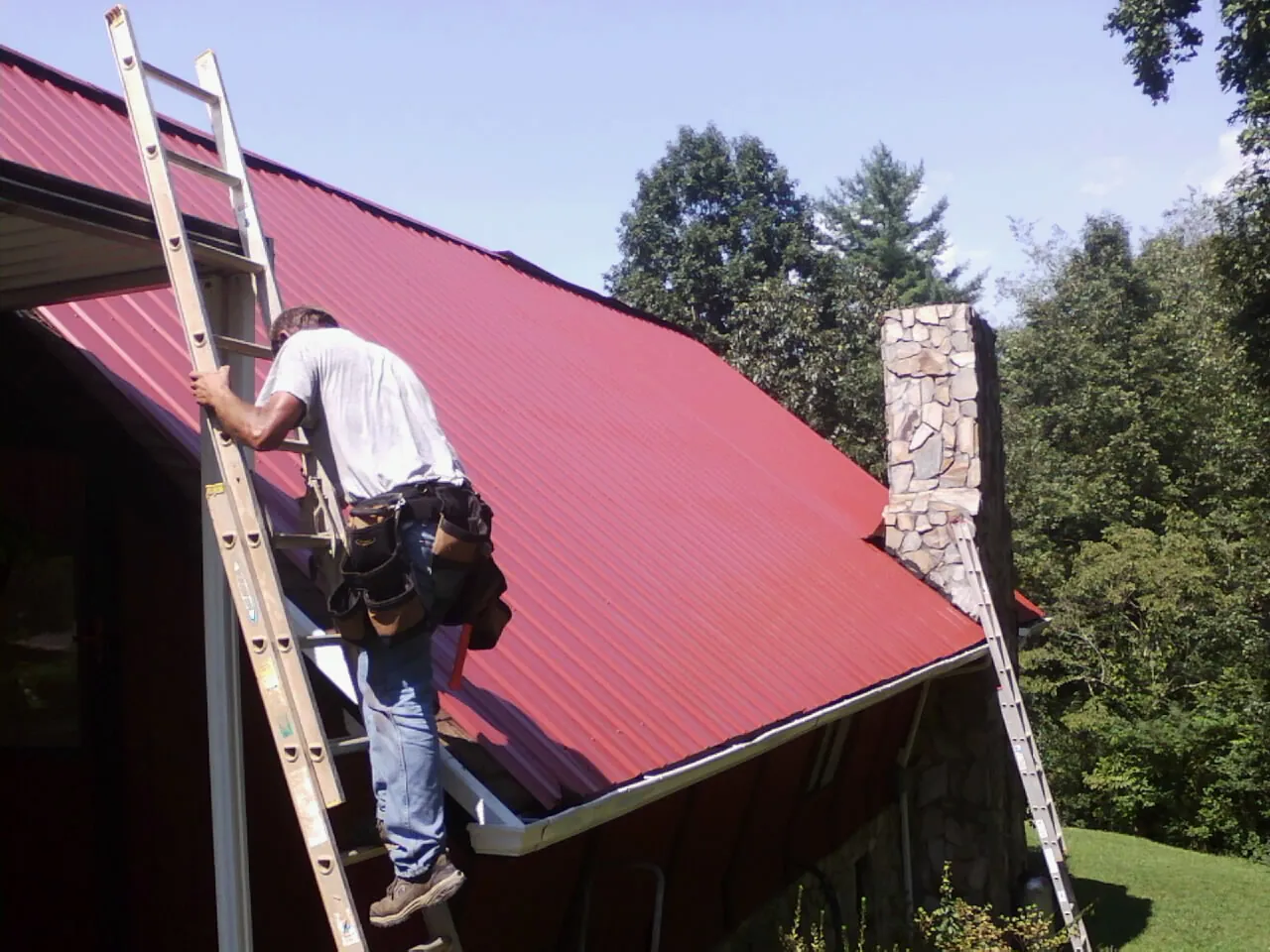 Roofer on a ladder beside a red metal roof and stone chimney — Soffit & Fascia Repair by Roofing Brothers in Saint John